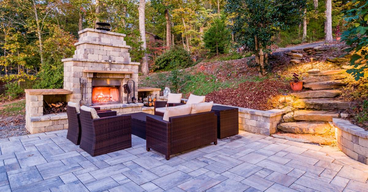 A stone outdoor fireplace with built-in side storage for chopped wood and logs accompanied by an outdoor couch and chairs.