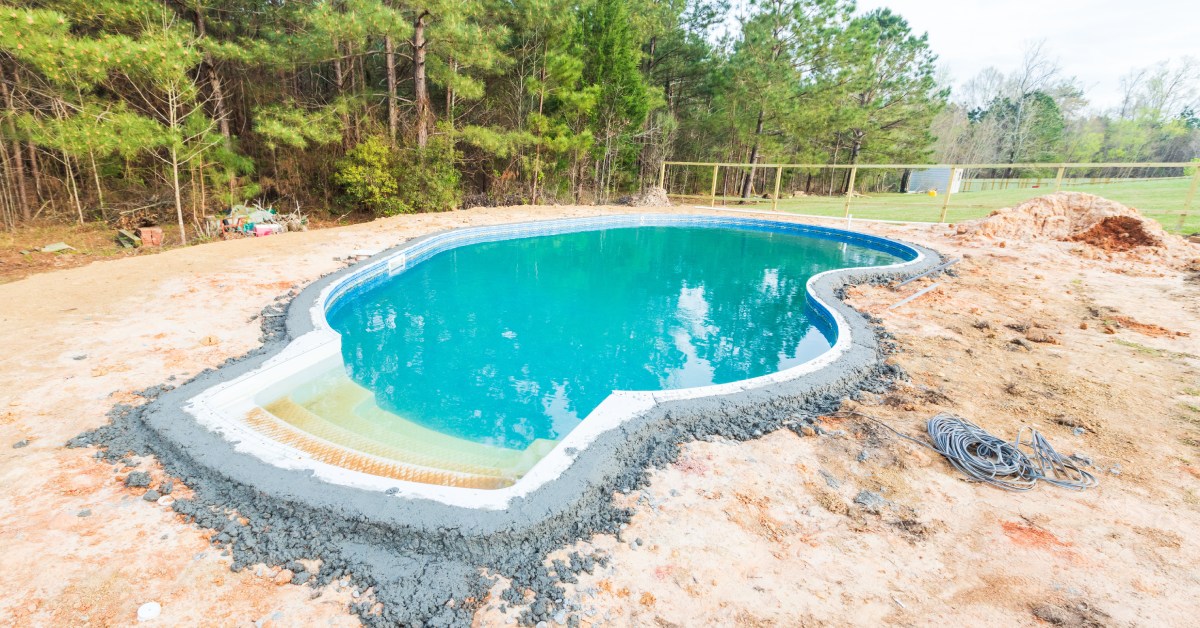 A yard with a swimming pool in the process of being constructed. There’s wet concrete and dirt surrounding it.