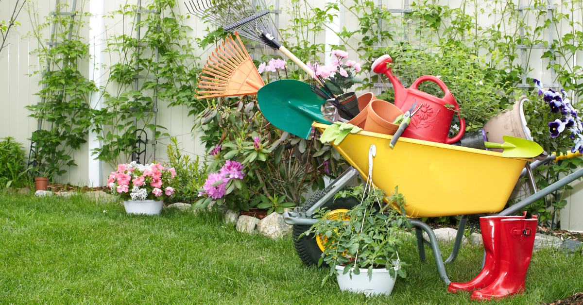 A wheelbarrow with various tools in it sitting in a yard next to a fence and a pair of red rubber boots.
