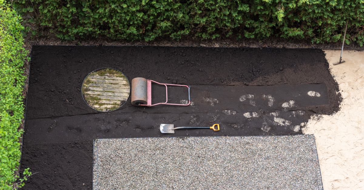 A yard surrounded by hedges in the process of being landscaped. There’s a roller and a shovel sitting on top of the dirt.