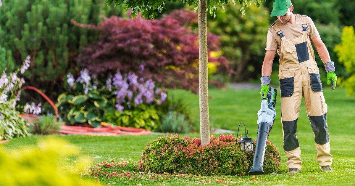 A man wearing light brown overalls and a green hat and gloves using a leaf blower to clean around a tree in a yard.