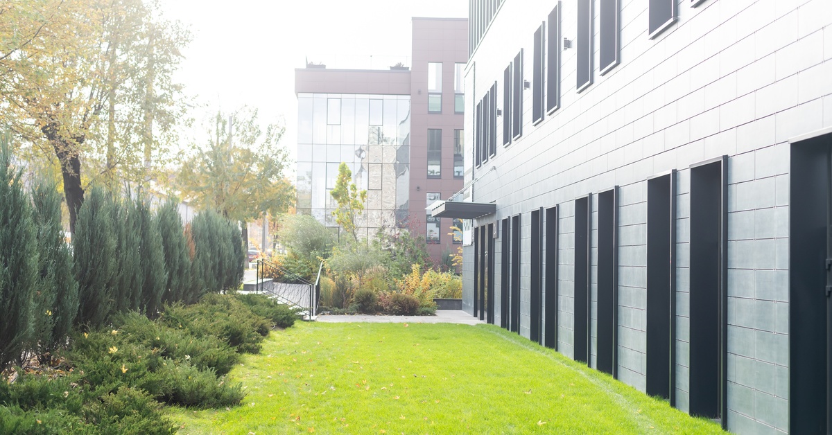 A bright green lawn next to a building with a row of trees and bushes on the other side beneath a hazy sky.