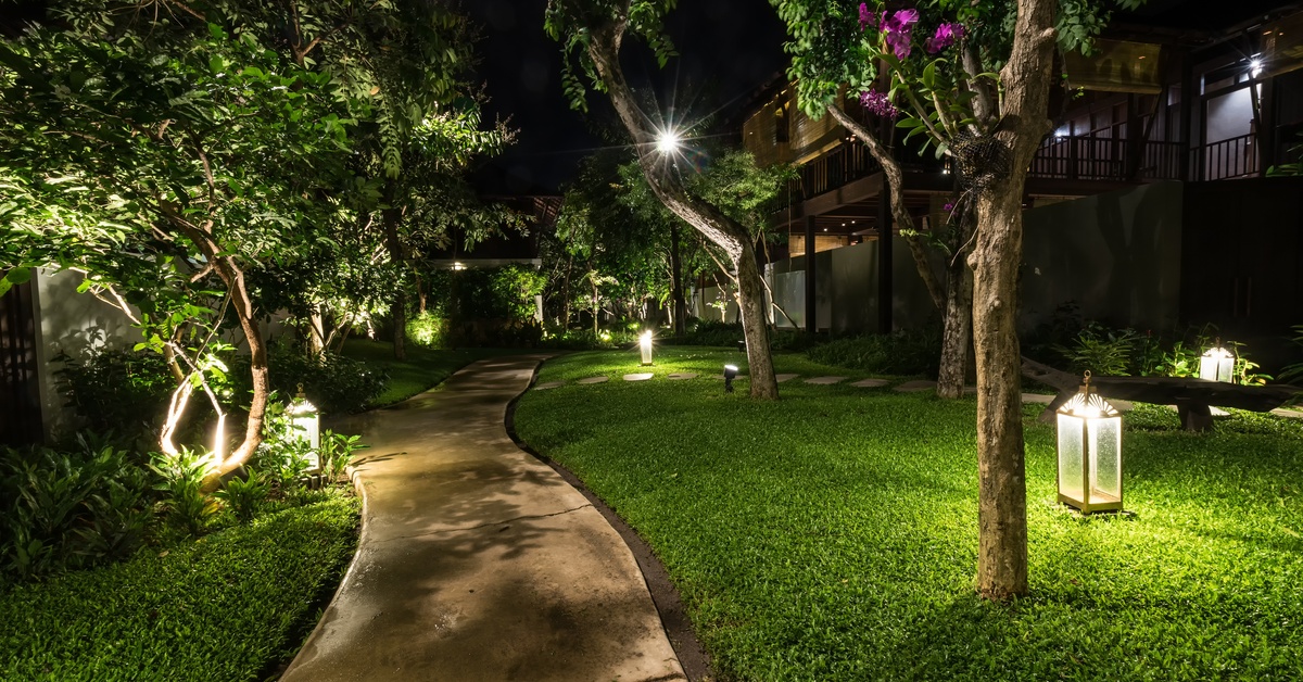 A concrete pathway in the backyard of a home features green grass, trees, and lights inside of glass lanterns.