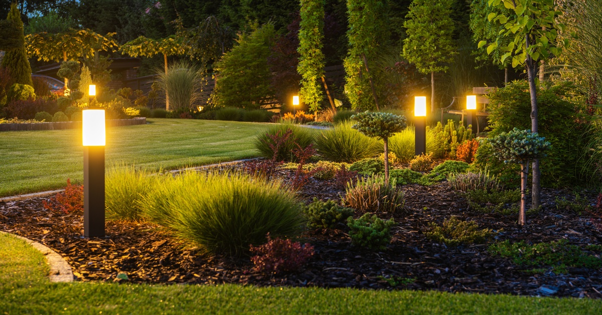 A lawn with short, green grass features trees, shrubs, and several short rectangular lights staked into the ground.