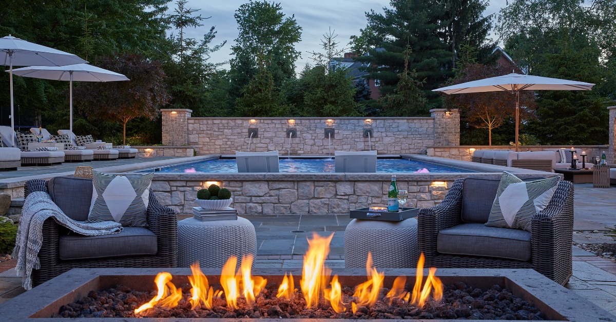 A patio with wicker chairs, umbrellas, a stone pool, wall lights, and flames burning in a rectangular fire pit.