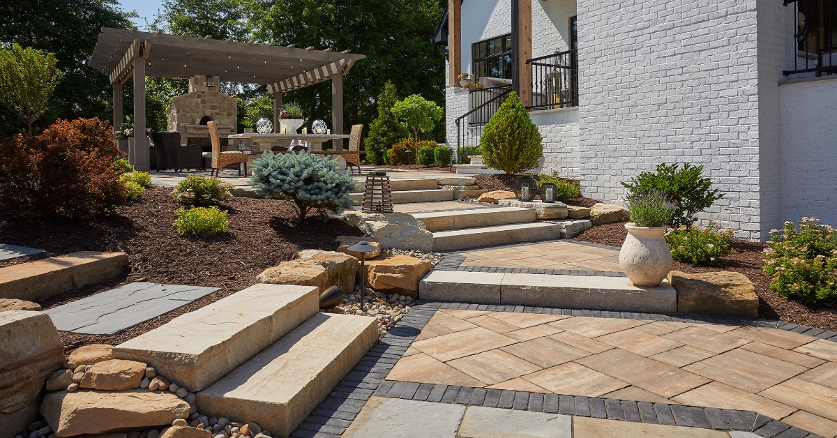 A paved garden walkway with stone steps, shrubs, a pergola, outdoor furniture, and a white brick house.