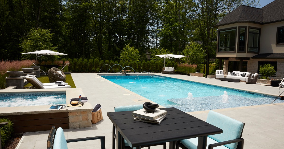 a backyard pool with lounge seating, patio umbrellas, water fountains, trees, and a large house beside it.