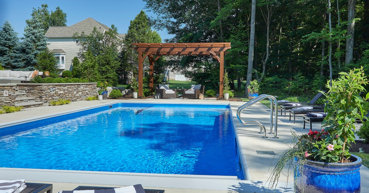 a blue backyard pool beside lounge chairs, a wooden pergola, stone wall, trees, and a large blue planter.