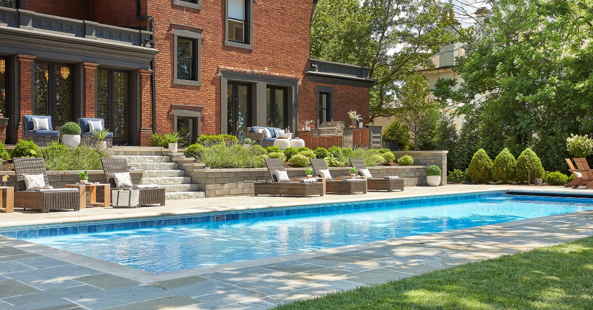 a backyard pool beside a brick house with lounge chairs, garden plants, stone steps, and a green lawn.