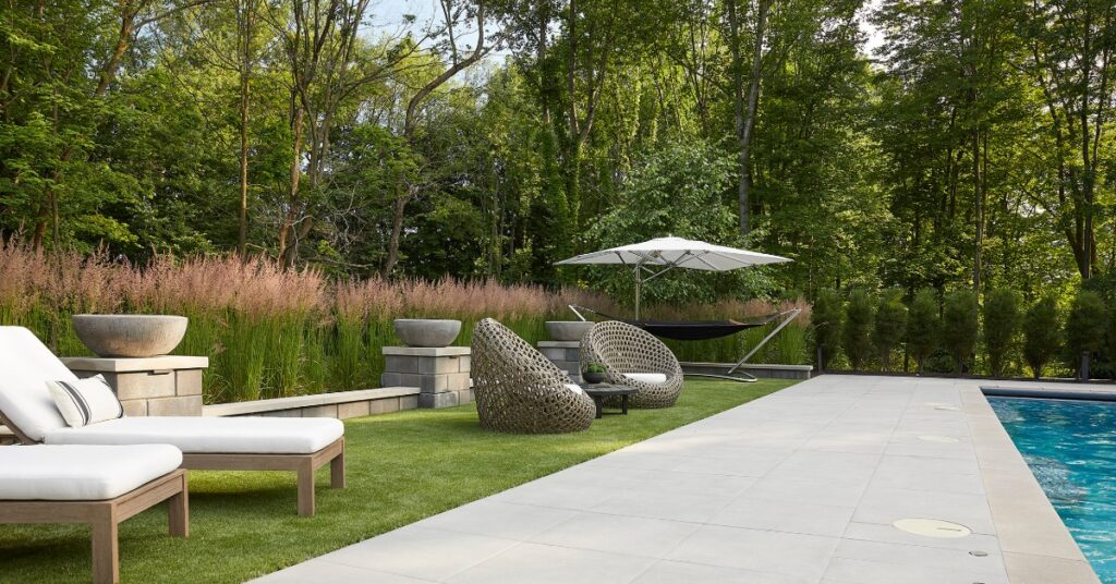 A backyard pool area with lounge chairs lined up along the grass. There are shrubs and tall trees behind the chairs.