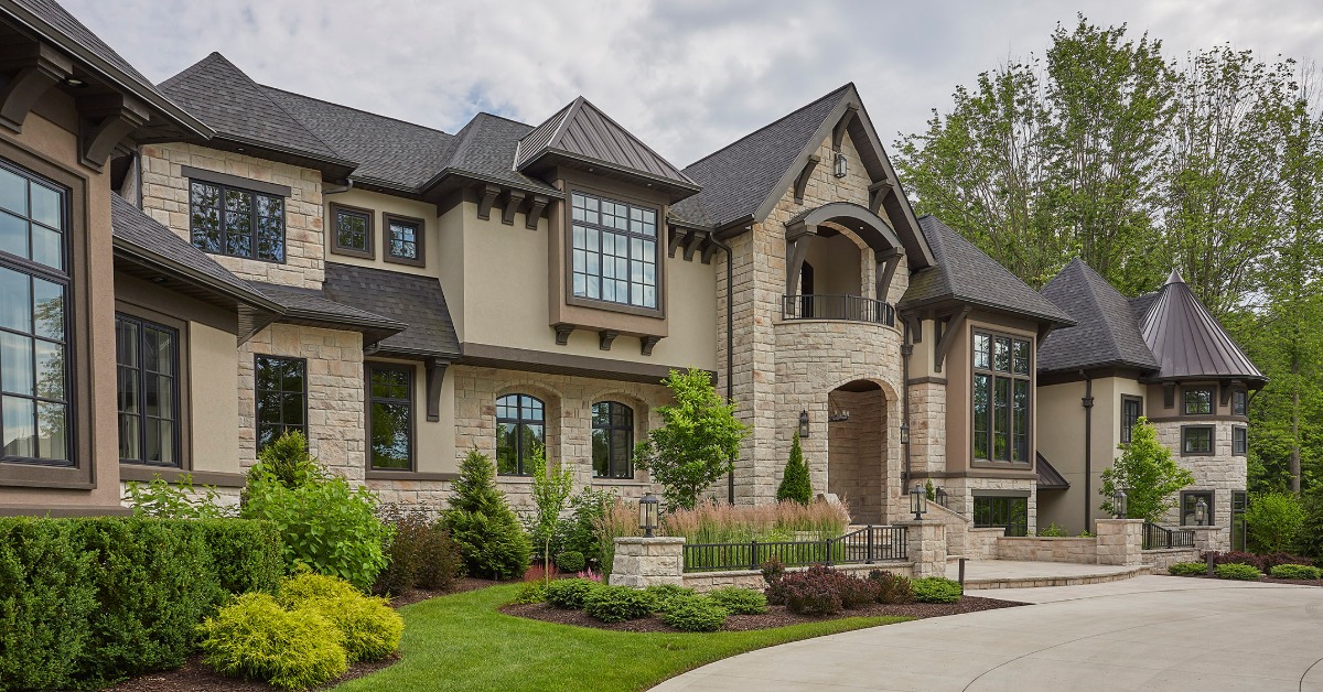 A large stone house with dark roofs and tall windows. Landscaped shrubs and small trees line the driveway.