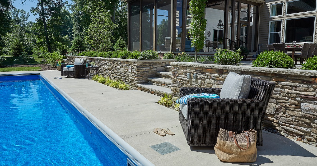 A backyard pool beside a retaining wall with small shrubs. The deck has chairs and steps leading to a covered porch.