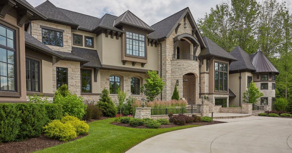 A large stone house with dark roofs and a well-maintained lawn. The lawn has shrubs and grass along the driveway.