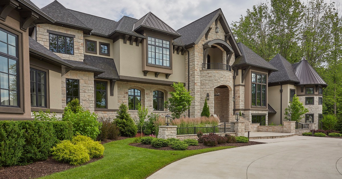 A large stone house with dark roofs and a well-maintained lawn. The lawn has shrubs and grass along the driveway.