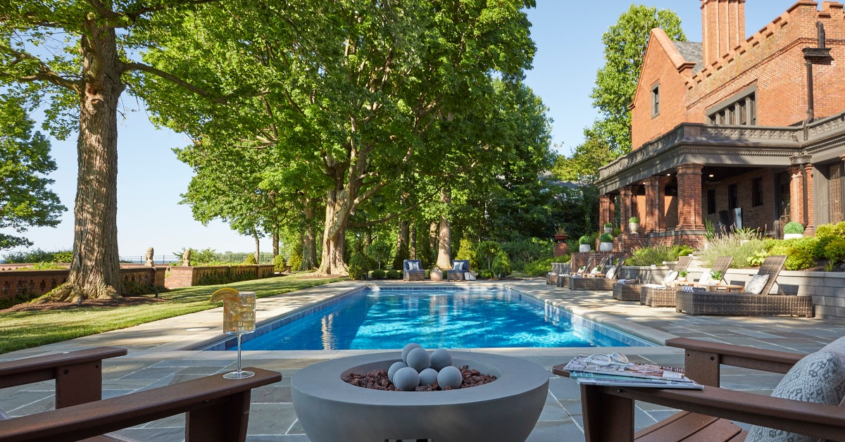 A backyard pool beside a brick house with lounge chairs, large trees, patio seating, and a round fire feature.