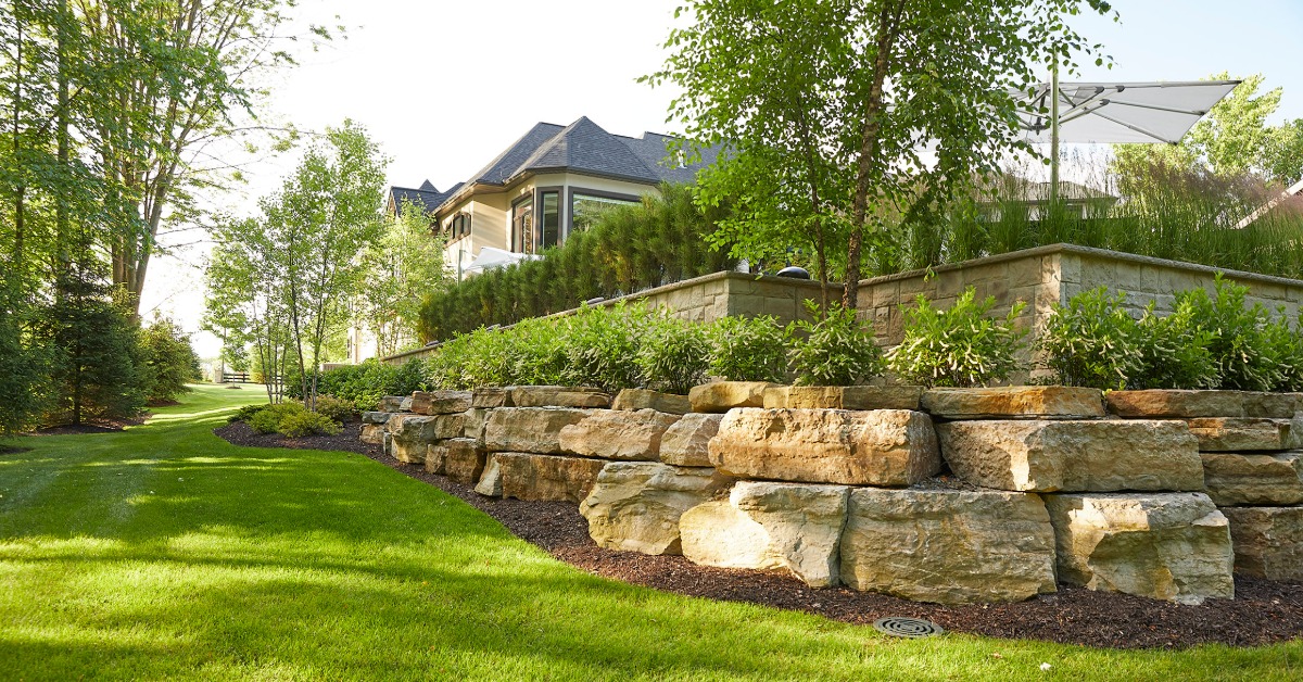 A landscaped yard with a stone retaining wall, green grass, shrubs, trees, and a house partly visible above.