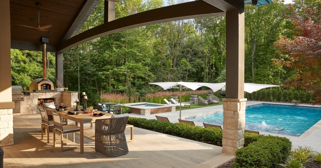 A covered patio with an outdoor dining table, pizza oven, swimming pool, lounge chairs, and trees beyond.