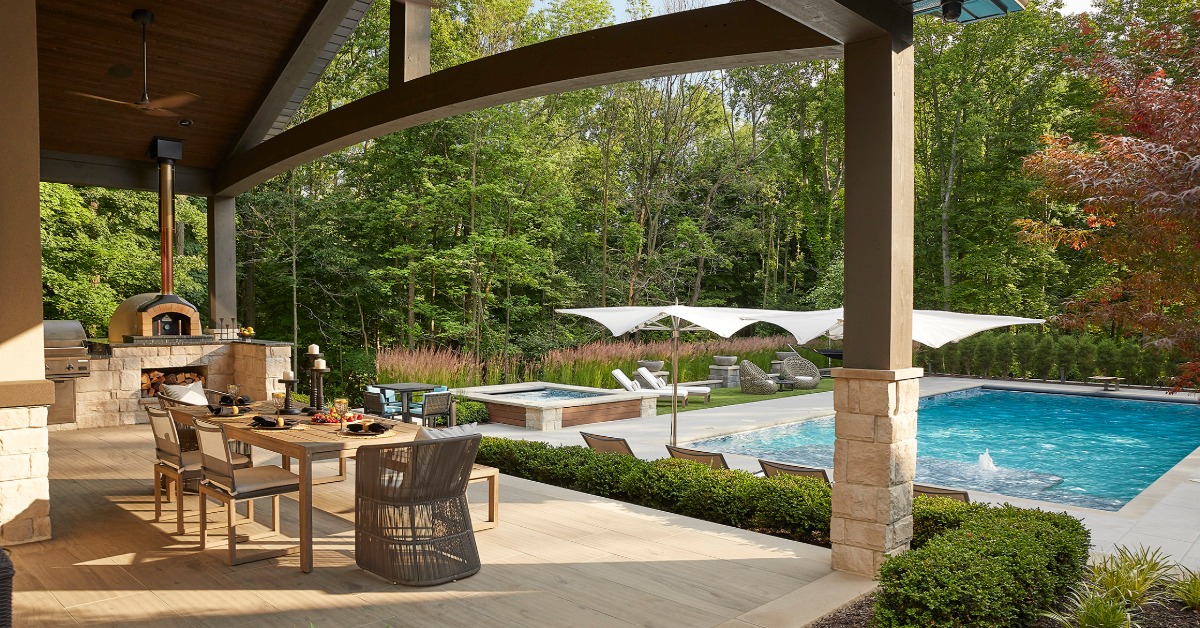 A covered patio with an outdoor dining table, pizza oven, swimming pool, lounge chairs, and trees beyond.