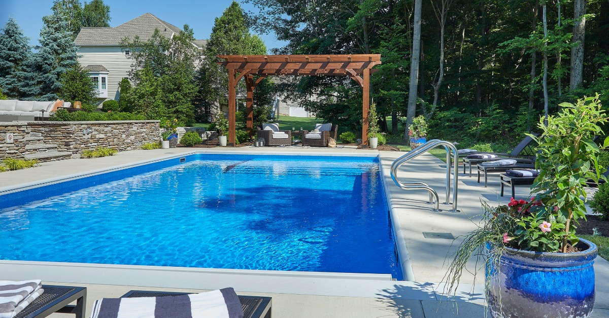 A rectangular swimming pool beside lounge chairs and stone walls. A wooden pergola stands along one side.