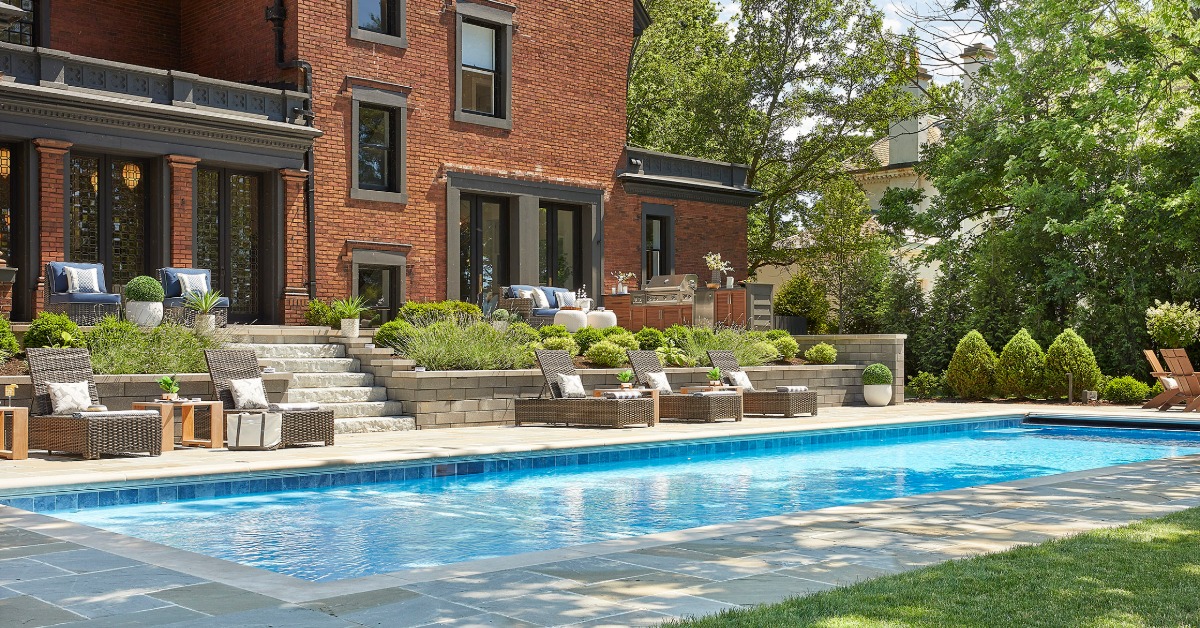 A long swimming pool behind a brick house, with lounge chairs, stone steps, shrubs, and patio seating.